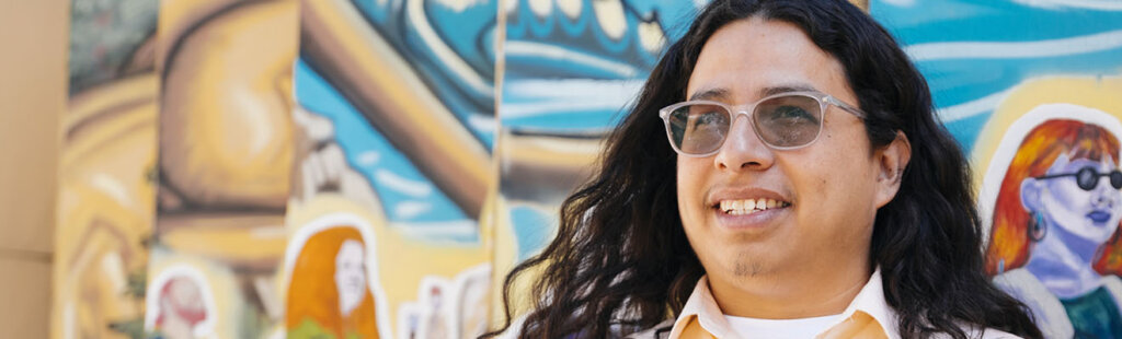 Person with long hair and glasses smiling in front of a colorful mural
