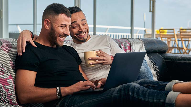 Male couple smiling while using a laptop sitting on the couch at home.