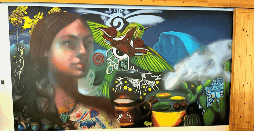 A large portrait of a woman stands surrounded by Native symbolism. Above her in gold is Coyolxauhqui, Aztec goddess of the moon. To her right, a hummingbird, or Copilli, represents a spiritual messenger and reminder of people’s Indigenous roots. To her left, a deer dancer appears beside a ceremonial cottonwood tree, and a small red handprint on her dress calls attention to the unreported sexual violence toward Indigenous women.