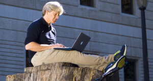 Person sitting on a tree stump working on a laptop.