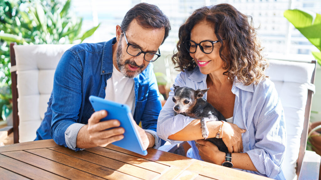Middle age hispanic couple using touchpad sitting on table with dog at terrace