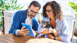 Middle age hispanic couple using touchpad sitting on table with dog at terrace