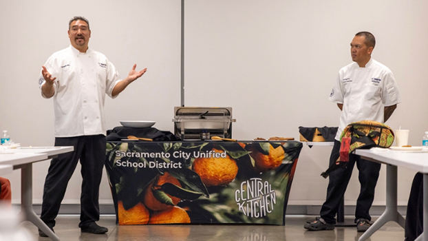 Two men in chefs jackets standing in front of a table