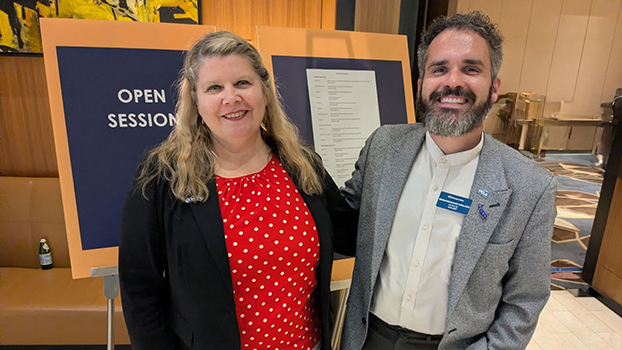 Two smiling people standing by the Regents "Open Session" sign