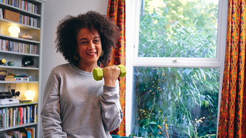 Woman lifting weights in her living room with a bookcase and window in the background