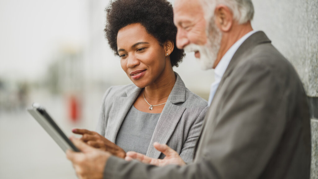 Shot of successful black businesswoman and her senior male colleague using digital tablet and having a discussion while standing against a wall of corporate building.