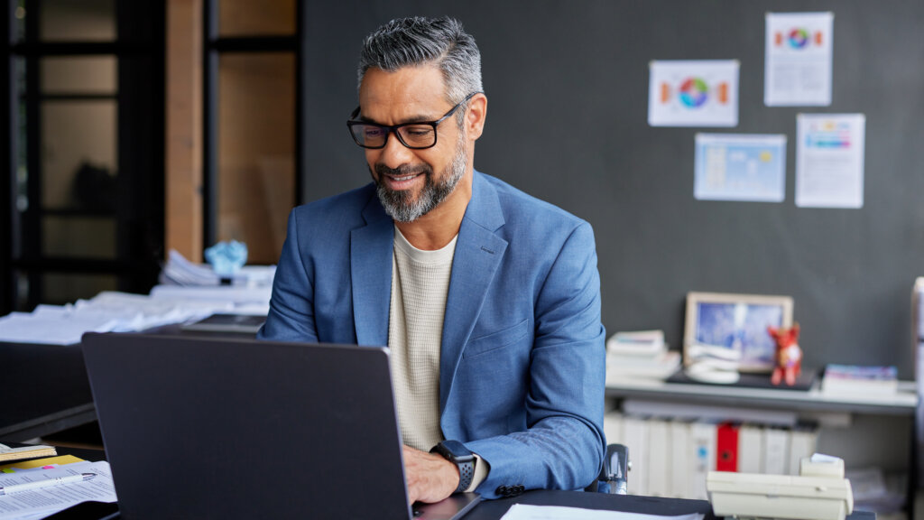 Mid adult businssman smiling while working on laptop in his office. Confident professional mixed race business man working in modern office while sitting at desk. Mature businessman engaged in online work while wearing eyeglasses.