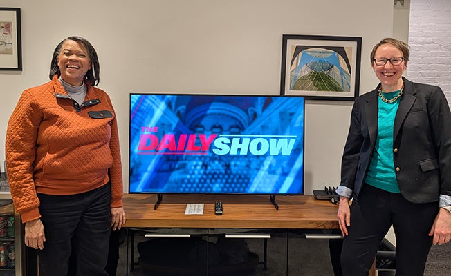 Mechele and Maura in front of a TV with "The Daily" show logo