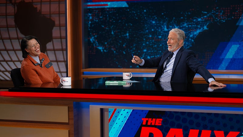 A. Mechele Dickerson and Jon Stewart laughing together on set at The Daily Show