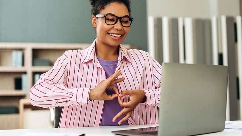 Happy young woman wearing glasses is using sign language while sitting at desk and having online conversation on laptop at office