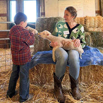 Hopland REC Community Educator Hannah Bird holds a lamb as a young visitor helps feed it during a field trip.