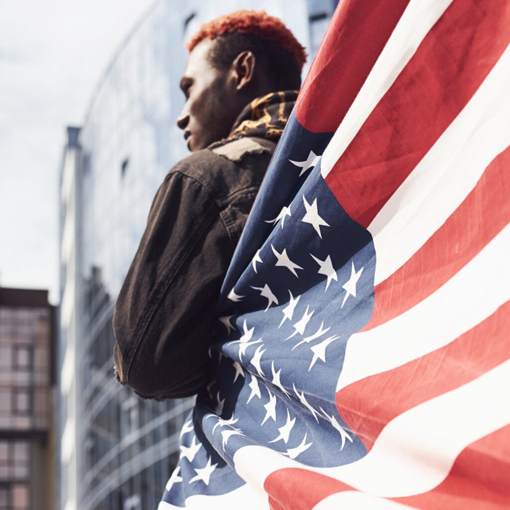 Young African American man in black jacket outdoors in the city standing against modern business building, partially wrapped in an American flag
