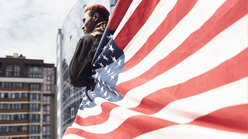 Young African American man in black jacket outdoors in the city standing against modern business building, partially wrapped in an American flag
