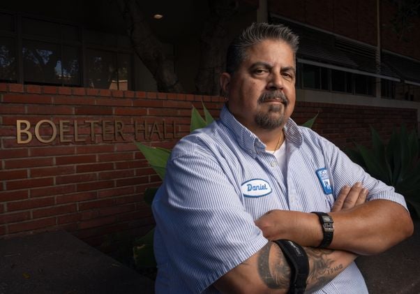 Daniel Estrada in janitorial uniform with arms folded
