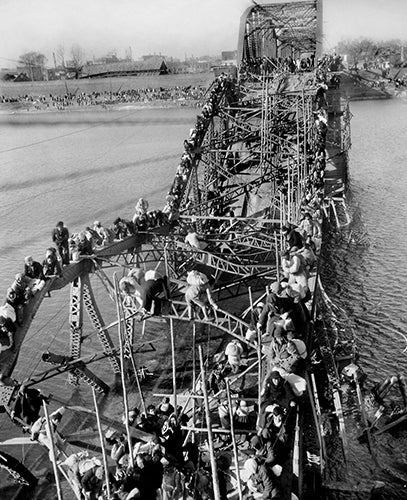 Huge number of people clinging to a dilapidated bridge above the water.