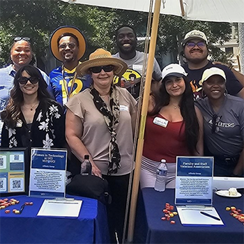 Group of smiling people standing at a picnic table under an umbrella