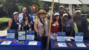 Group of smiling people standing at a picnic table under an umbrella